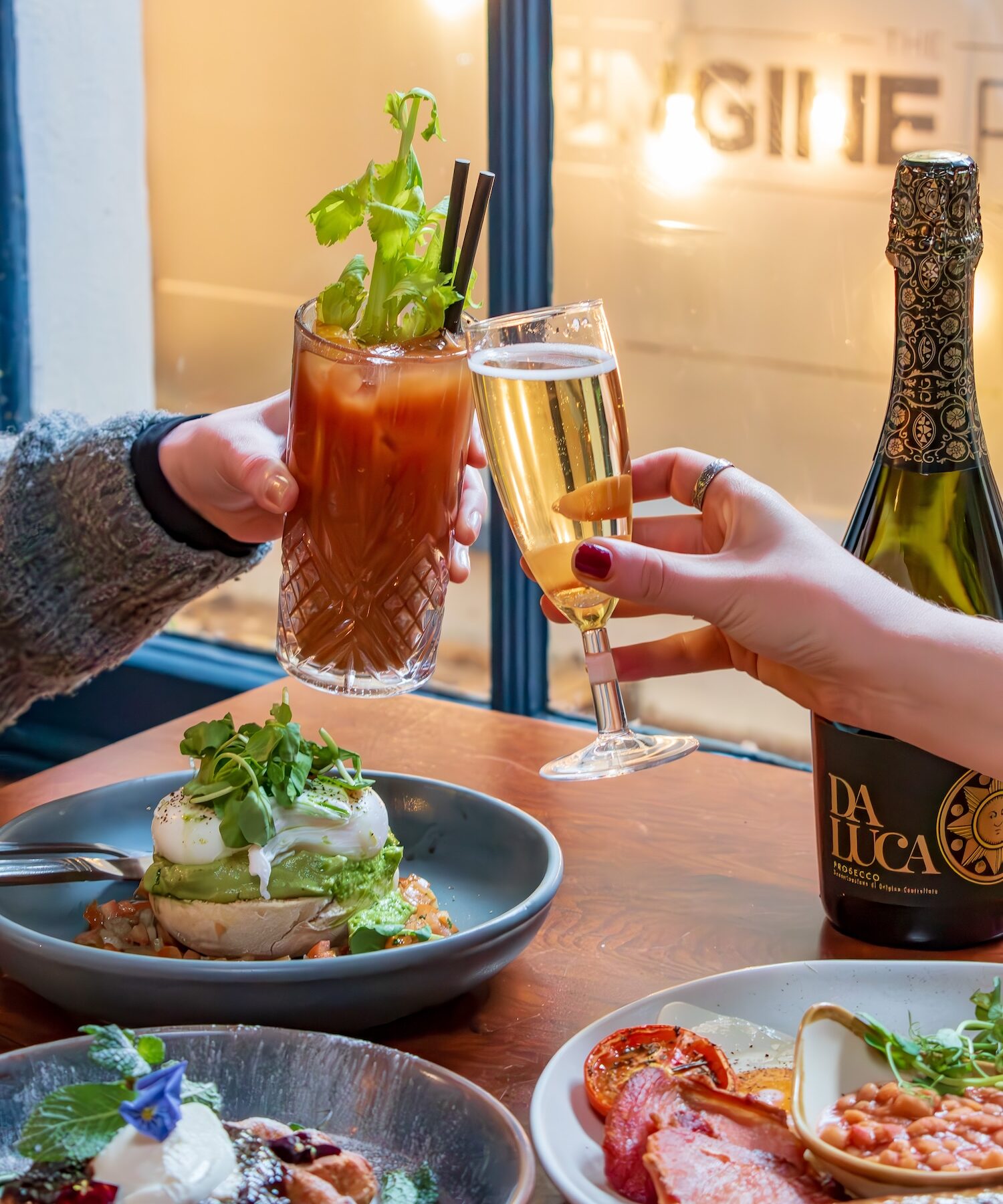 A toast to the best bottomless brunch at Racks Bar & Kitchen in Bristol. Two hands clink glasses of Prosecco and Bloody Mary over a table laden with brunch classics, including avocado toast and a traditional full English breakfast. This image exudes the joy of a vibrant bottomless brunch in a cosy Bristol venue.